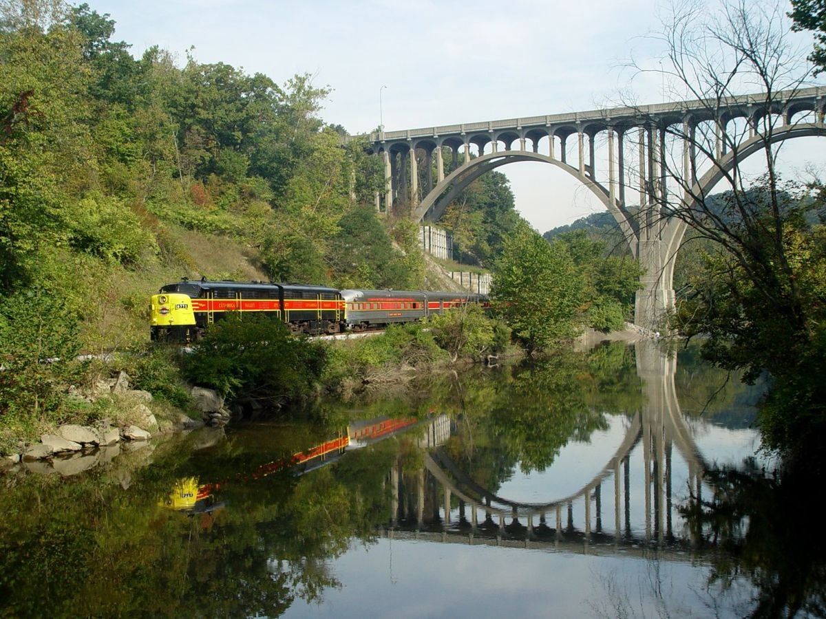 The sleek-looking train of the Cuyahoga Scenic Valley Railroad traverses under a majestic bridge, past glassy water, and through lush foliage.