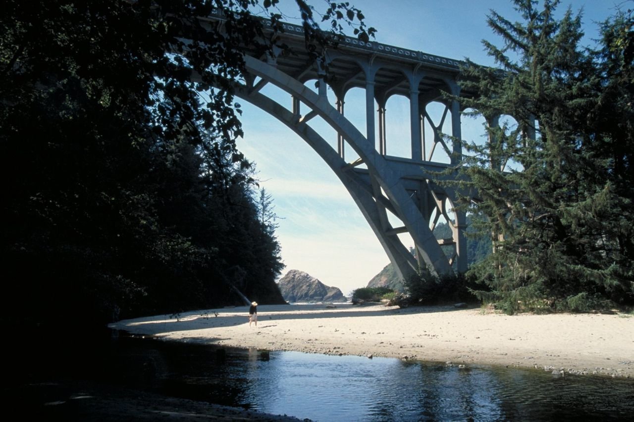 A visitor strolls on a sandy beach beneath an arch bridge on Oregon's Pacific Coast Scenic Byway.