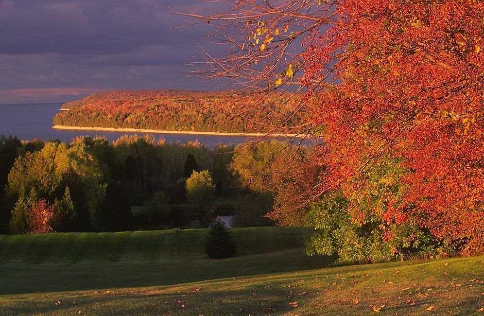 Grandview Overlook rests atop the Niagara Escarpment bluffs of Ellison Bay, Wisconsin looking northward. The scarlet and bittersweet autumn foliage is interspersed with Brunswick-green cedar trees. The foliage, Kelly-green slopes and golden shorelines all provide a spectacular contrast against backdrops of darkening periwinklepink skies and the ultramarine waters of Green Bay near sunset.